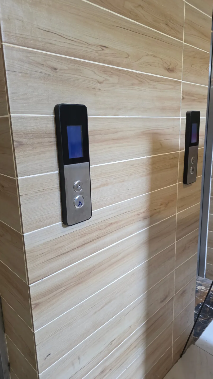 Two black-framed TFT elevator landing panels with blue colour displays and stainless call buttons installed on a wood-grain tiled lift lobby wall in Lahore