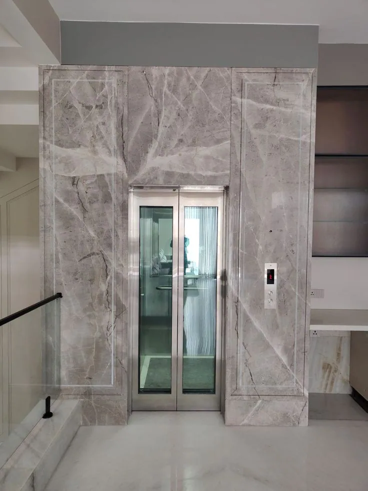 Stainless steel home elevator landing door with full-height laminated glass vision panels set in a marble-clad residential portal