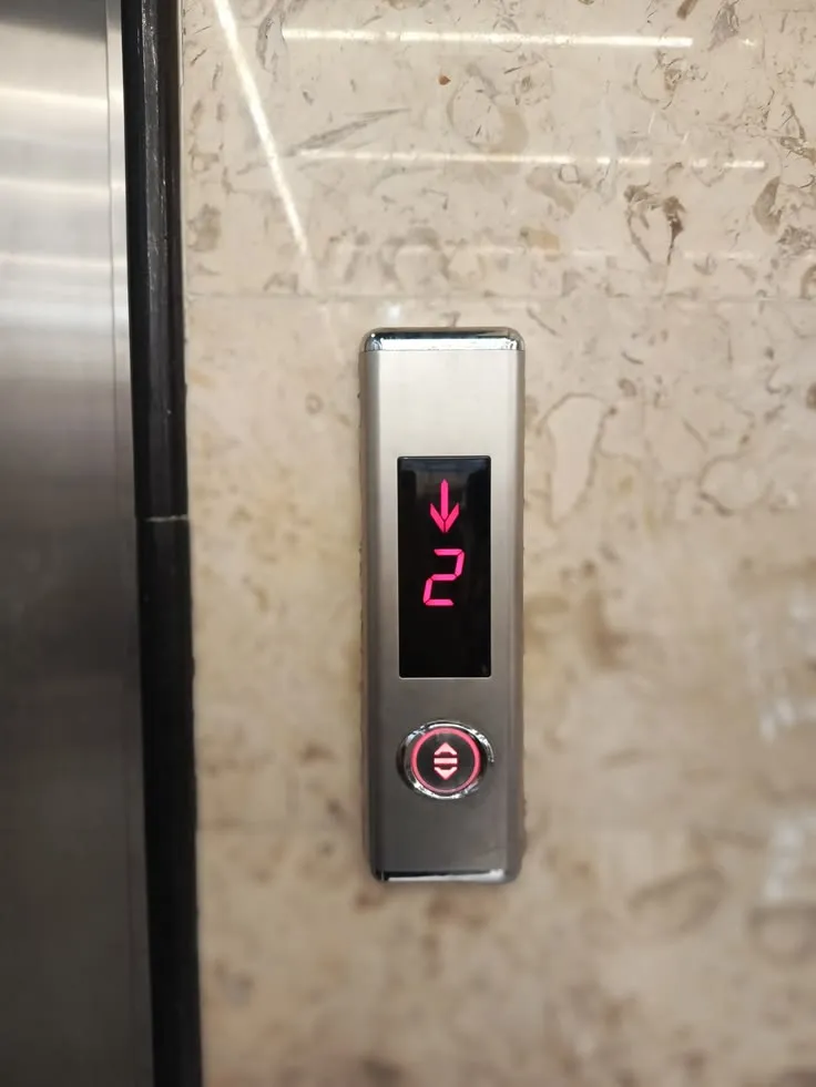 Slim stainless steel elevator landing operating panel with red LED position indicator, down direction arrow and round illuminated call button on marble lobby wall