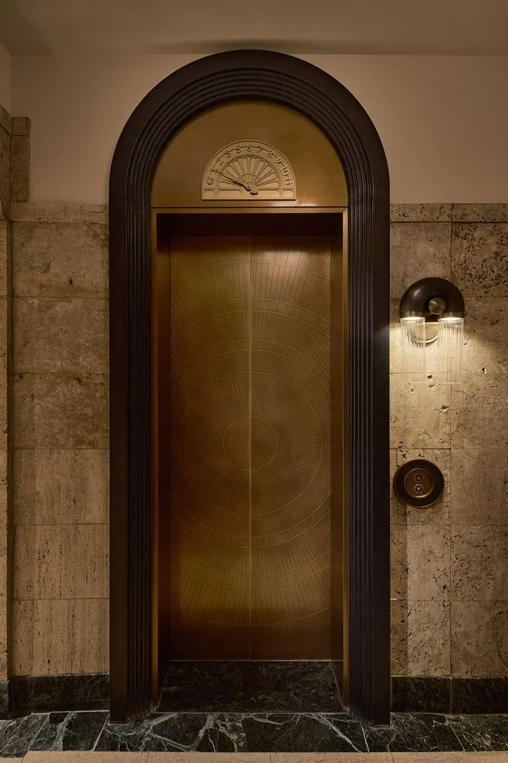 Antique brass elevator landing door with etched sunburst pattern and vintage arc floor indicator above the header, hotel lobby installation