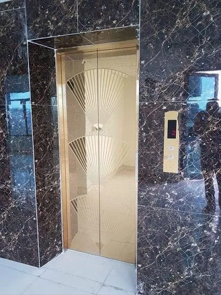 Gold titanium elevator door with perforated fan pattern and marble portal cladding, supplied and installed in Karachi